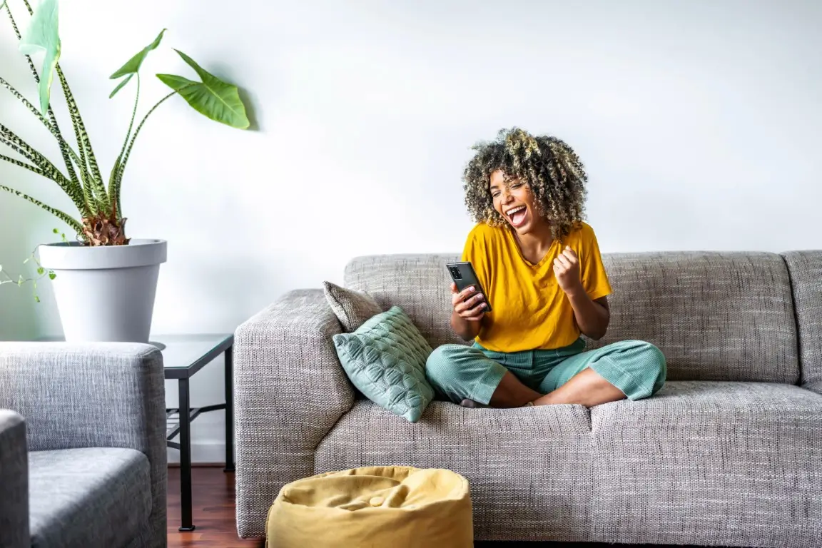 Excited happy young black woman holding smart phone device sitting on sofa at home.